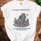 White t-shirt with graphic of a black cat gaming, holding controller, with text "We Must Remain Silly To Survive The Horrors Of This World."