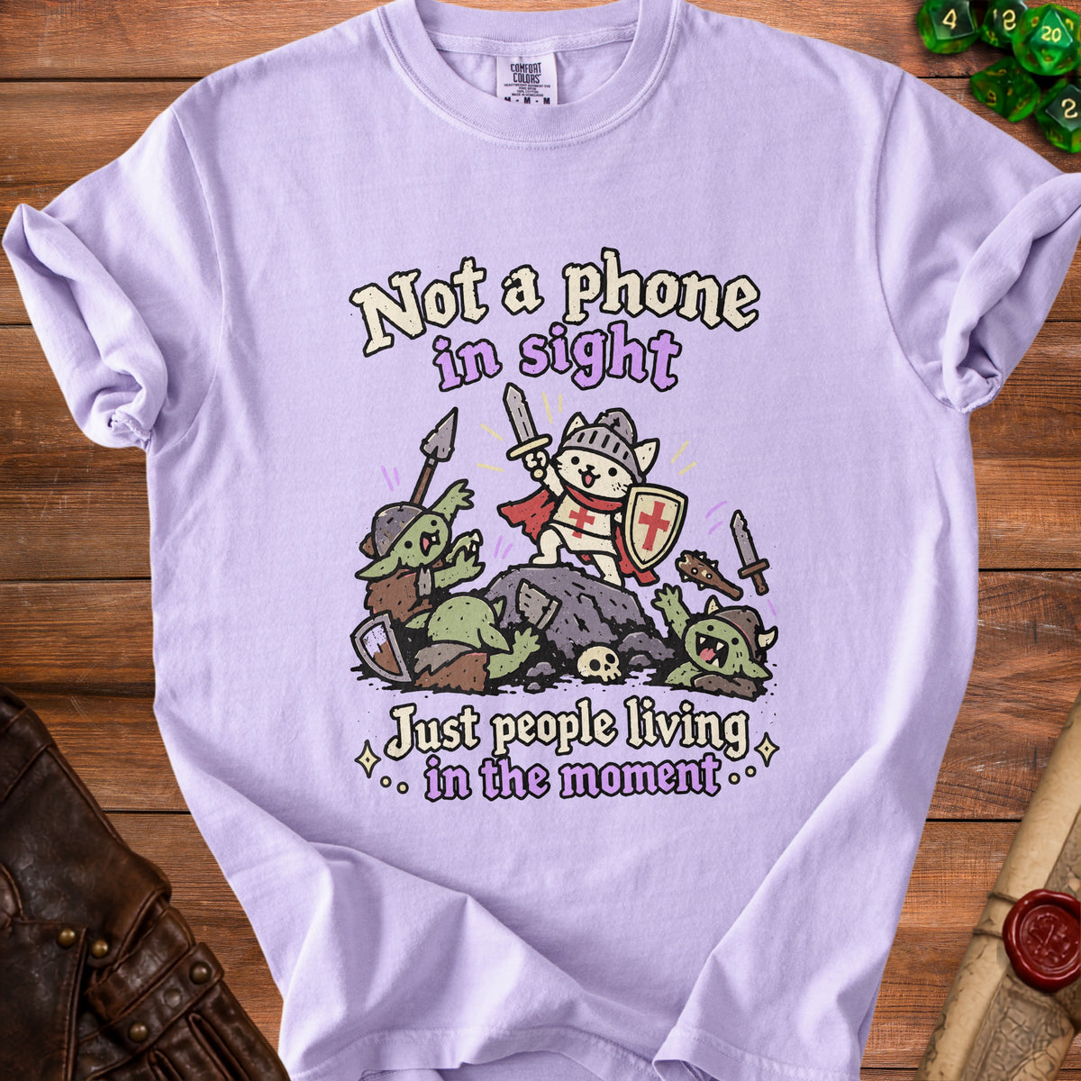Purple t-shirt graphic with cat knight fighting goblins, text "Not a phone in sight. Just people living in the moment."