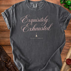 Dark gray t-shirt with pink text reading "Exquisitely Exhausted" and "the curse is adulthood".