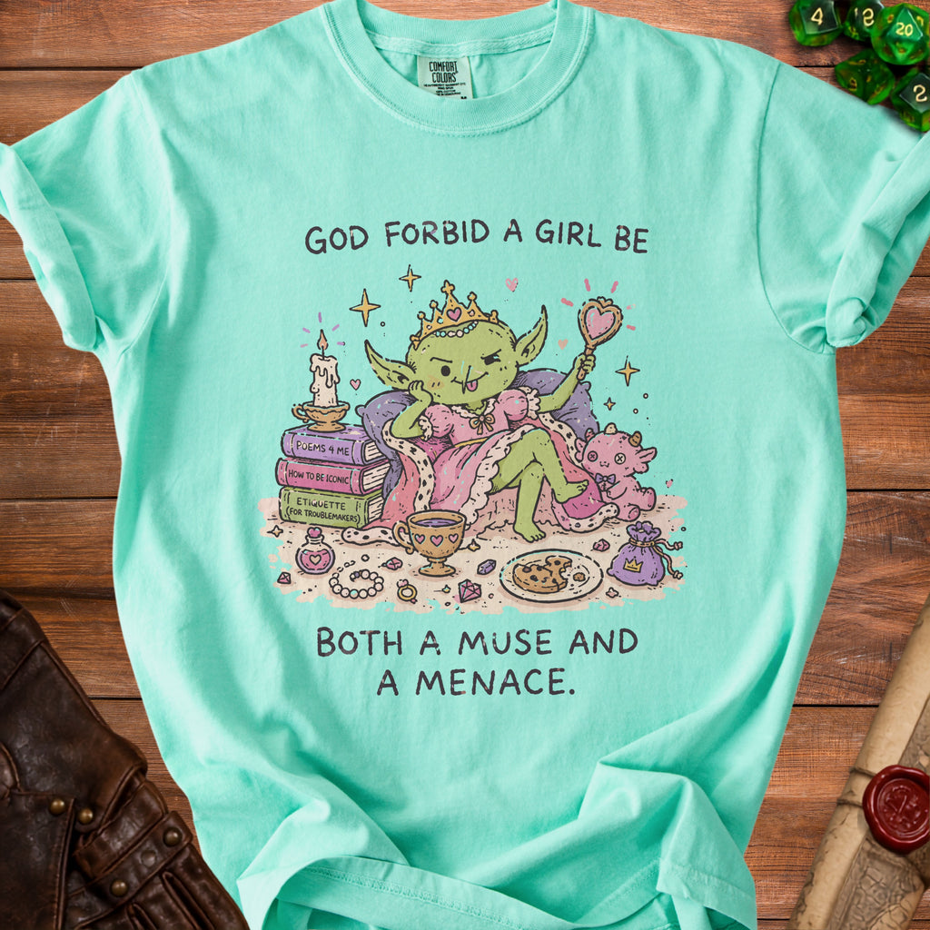 Green t-shirt with cartoon goblin holding mirror, text says "God forbid a girl be both a muse and a menace."