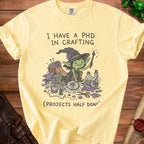 Yellow t-shirt with a graphic of a witch surrounded by crafting supplies and text.