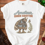 White t-shirt with graphic of moth holding binoculars and text "Amateur Omen Spotter".