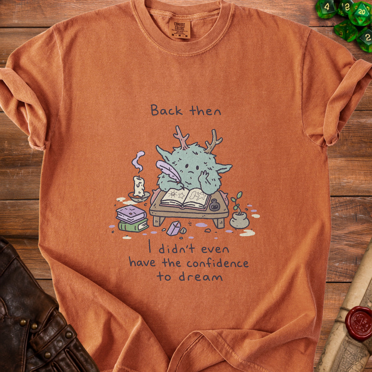 Orange t-shirt with cartoon creature writing and text "Back then I didn't even have the confidence to dream."
