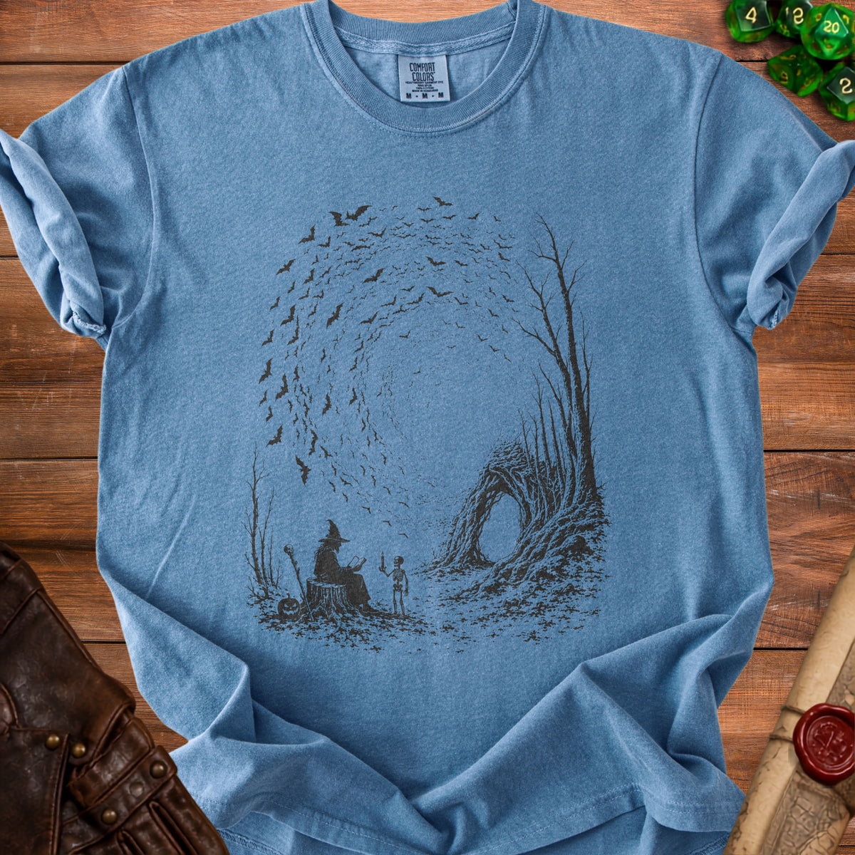 Blue t-shirt with a dark graphic of a witch, skeleton, and bats in a forest.