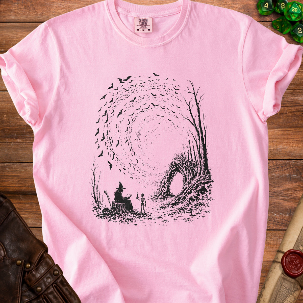 Pink t-shirt with a black graphic of a witch, skeleton, bats, and trees.