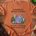 Orange t-shirt with "Currently Being Cursed By The Universe (And Deadlines)" text.