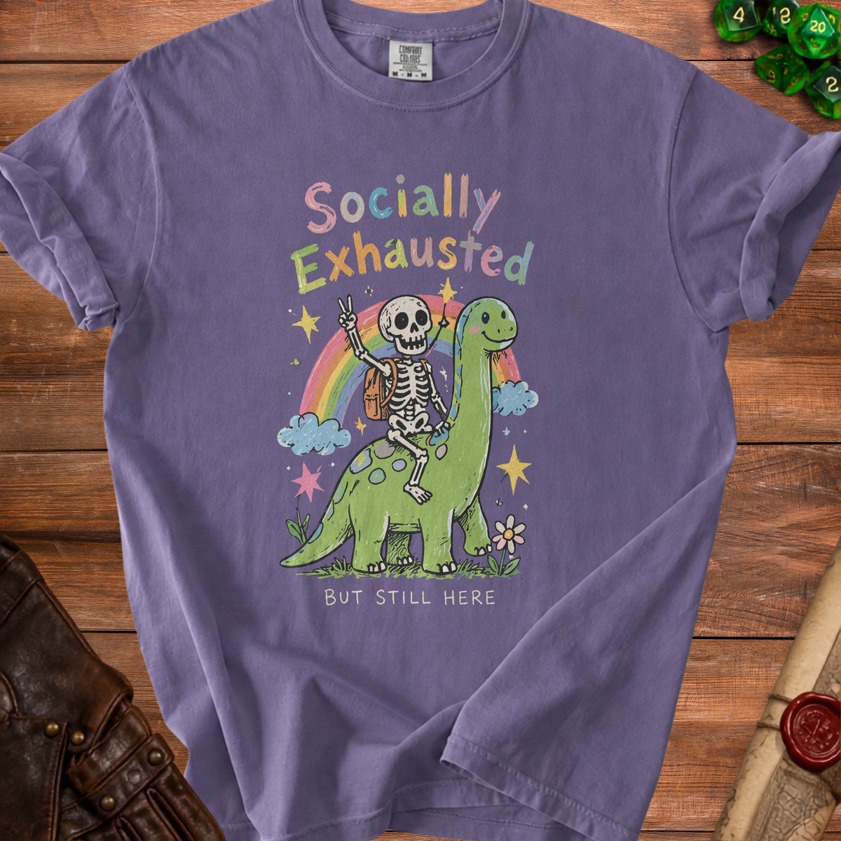 Purple t-shirt with skeleton riding dinosaur, rainbow, and text "Socially Exhausted But Still Here".