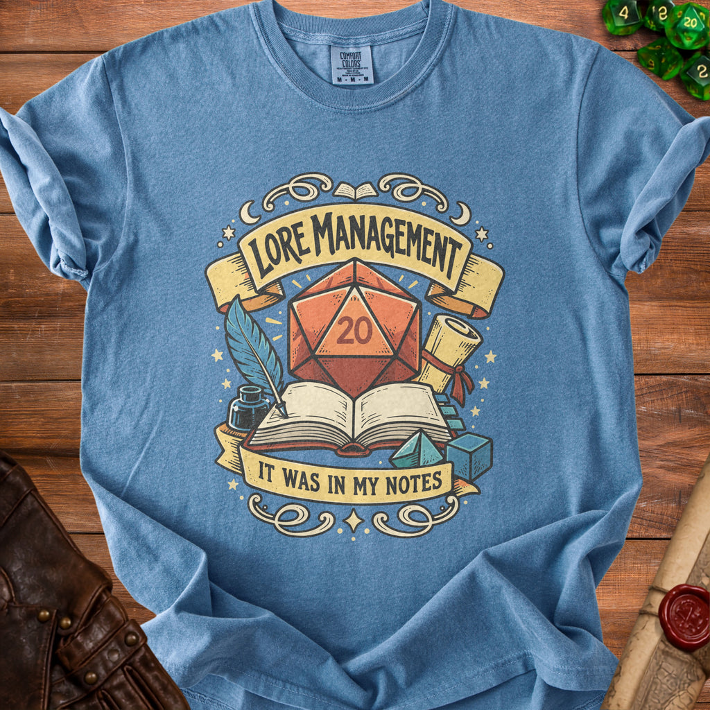 Blue t-shirt with dice, book, quill, and text "LORE MANAGEMENT IT WAS IN MY NOTES".