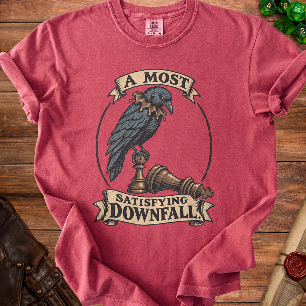 A red t-shirt printed with a crow wearing a ruff collar, perched on a chess piece, and text reading A Most Satisfying Downfall.