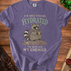 Purple t-shirt graphic: raccoon wearing cape drinks from goblet, text "I'm only staying hydrated to outlive my enemies".
