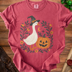 Goose wearing witch hat with pumpkin and floral wreath on pink t-shirt.