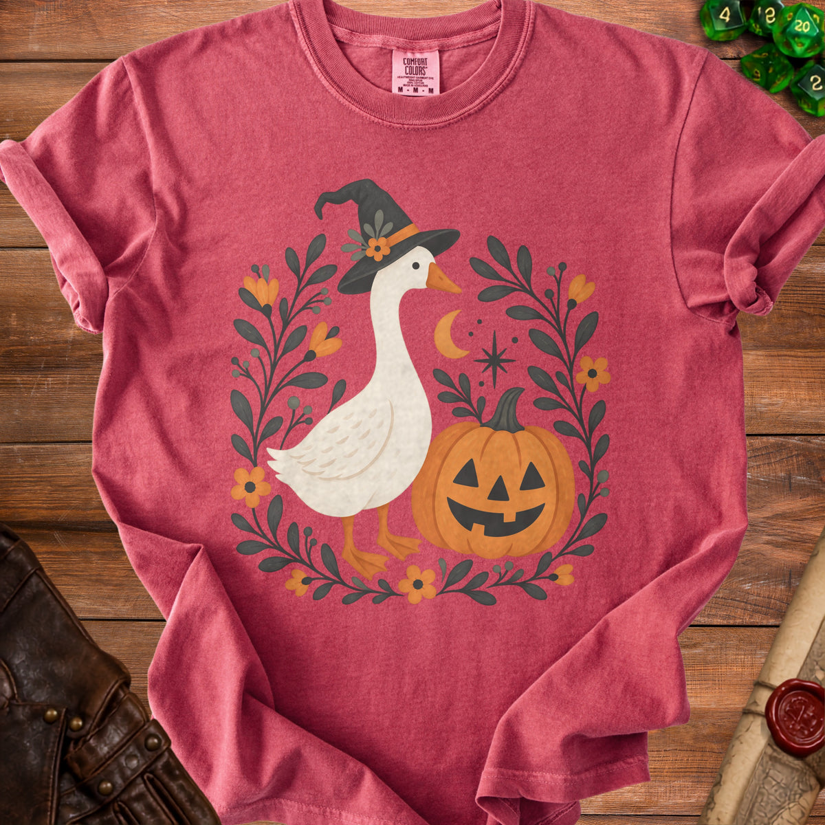 Goose wearing witch hat with pumpkin and floral wreath on pink t-shirt.