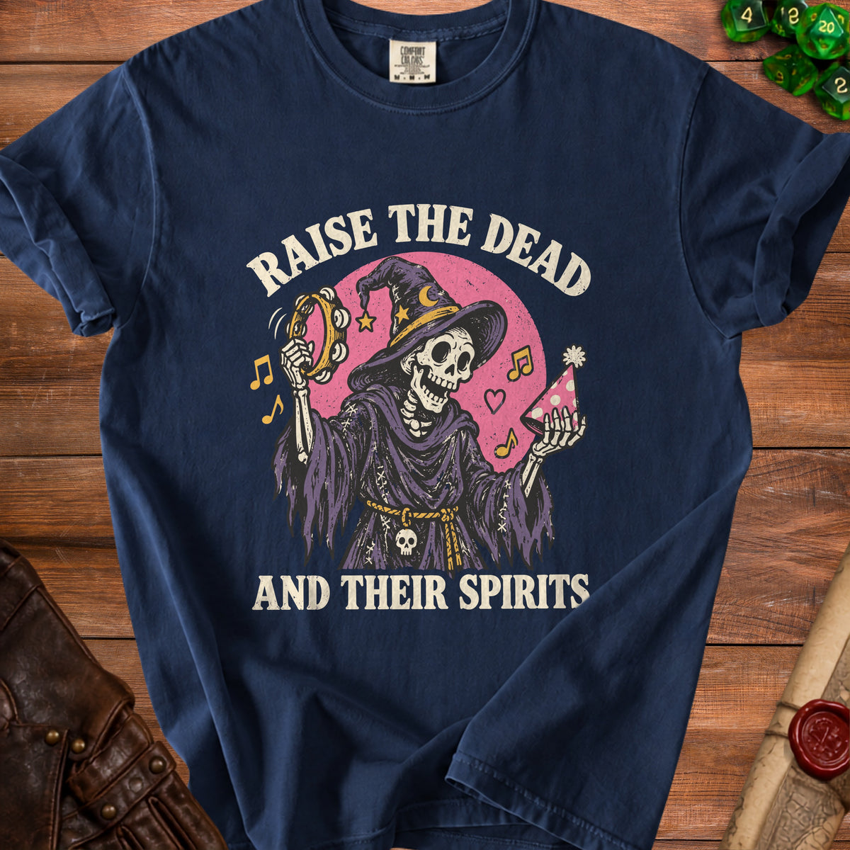 A dark blue t-shirt with a skeleton in a witch hat holding a tambourine and party hat, surrounded by the text RAISE THE DEAD AND THEIR SPIRITS.