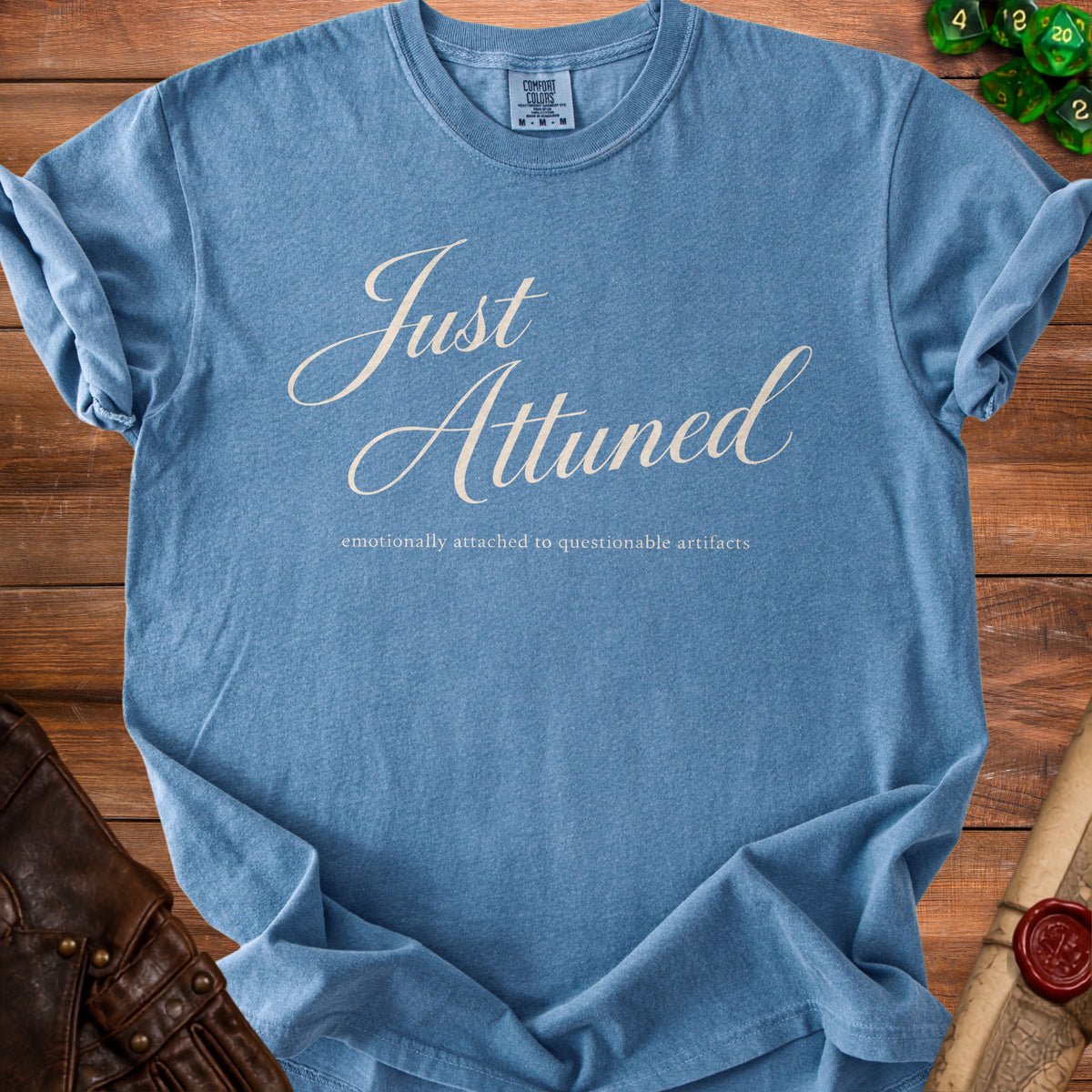 Blue t-shirt featuring the text Just Attuned and emotionally attached to questionable artifacts, laid on wood.