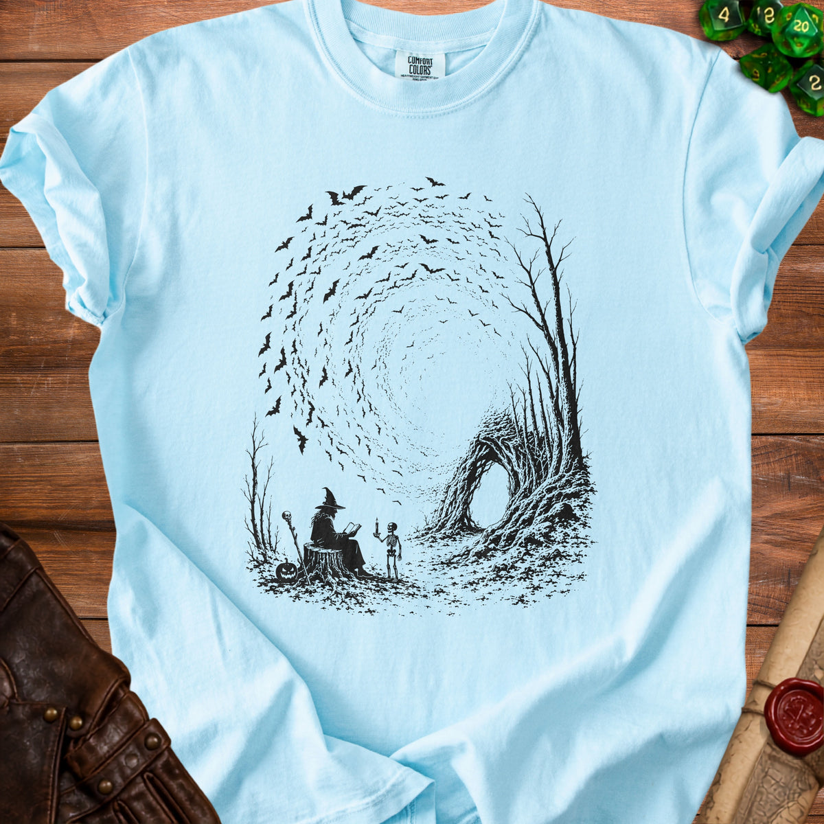 Black ink drawing on light blue t-shirt of a witch reading to a skeleton surrounded by bats.