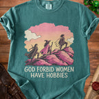 Teal t-shirt featuring an illustration of three female adventurers climbing a rocky hill, with text reading GOD FORBID WOMEN HAVE HOBBIES.