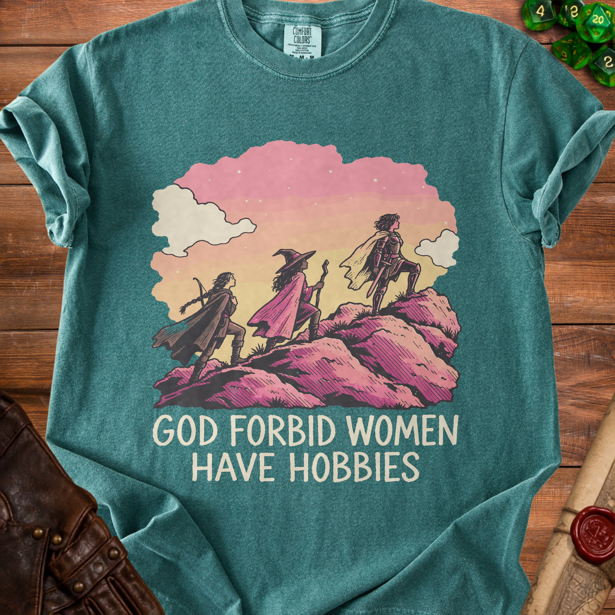 Teal t-shirt featuring an illustration of three female adventurers climbing a rocky hill, with text reading GOD FORBID WOMEN HAVE HOBBIES.
