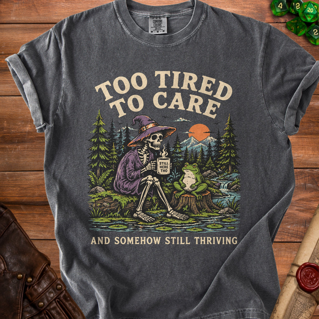 Dark grey t-shirt graphic: skeleton with mug, frog, and forest scene. Text: "Too Tired to Care" and "And Somehow Still Thriving".