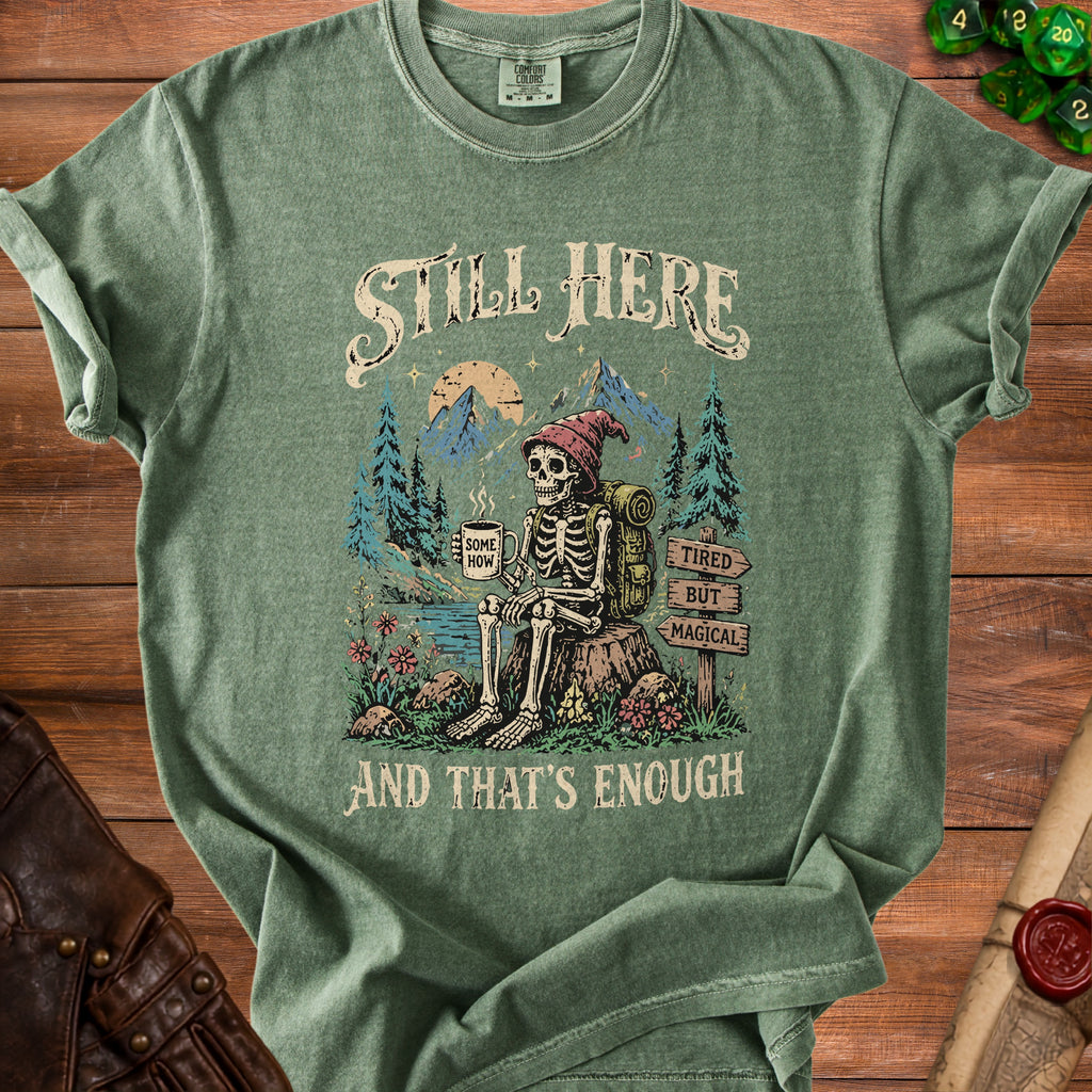 Green t-shirt with a skeleton sitting with a mug, mountains, and trees. Text reads "Still Here".