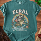 T-shirt graphic with text "FERAL but in a whimsical way" featuring an opossum riding a frog.