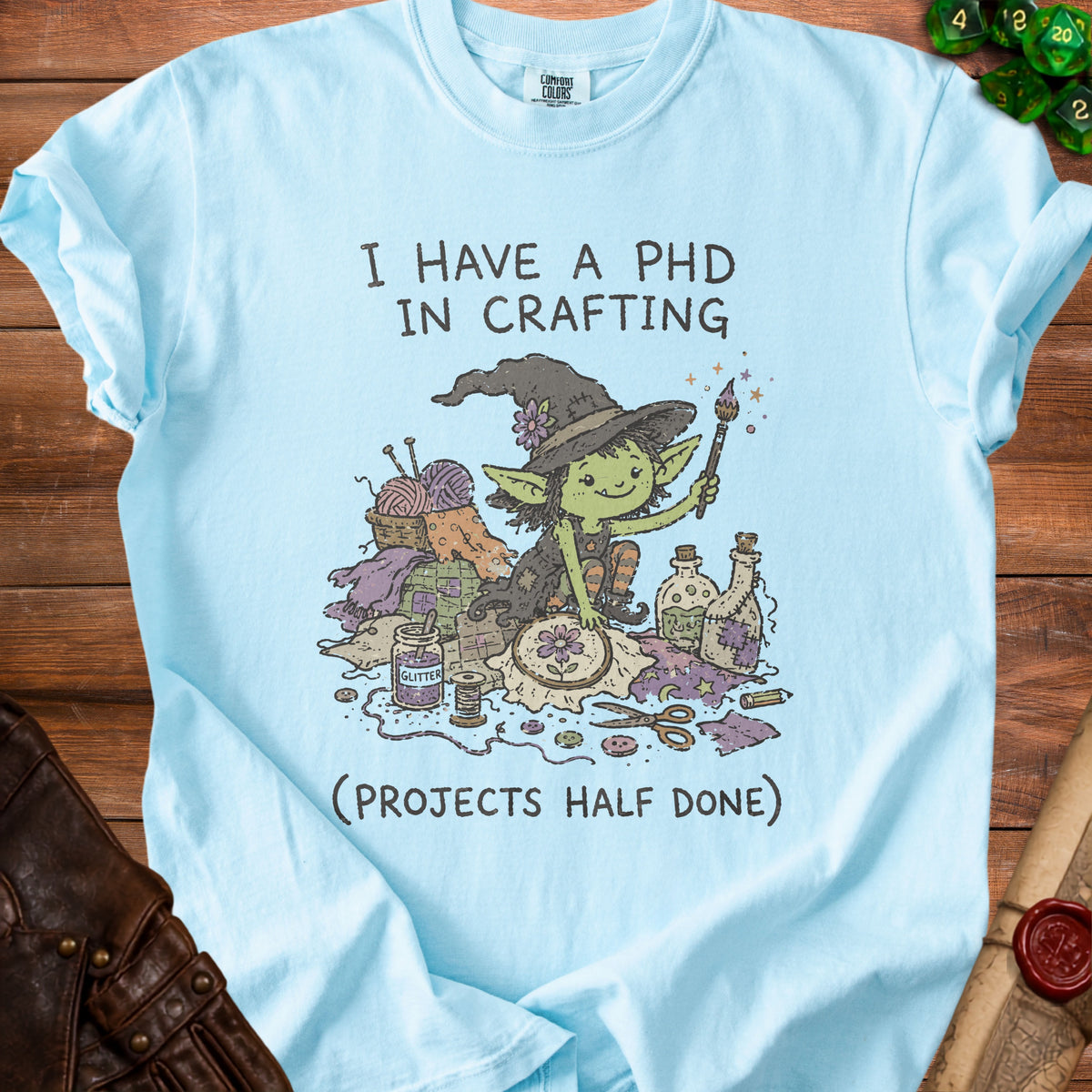 Light blue t-shirt with graphic of a crafting goblin and text "I HAVE A PHD IN CRAFTING (PROJECTS HALF DONE)".