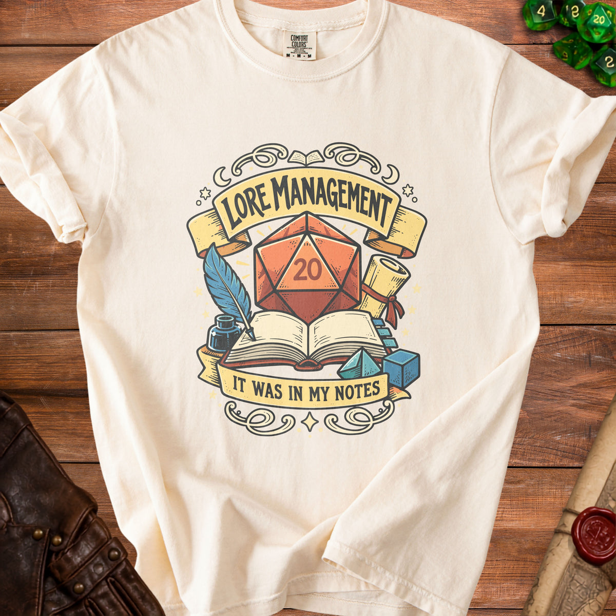 Cream t-shirt with "Lore Management" text, d20 die, open book, quill, and inkwell.