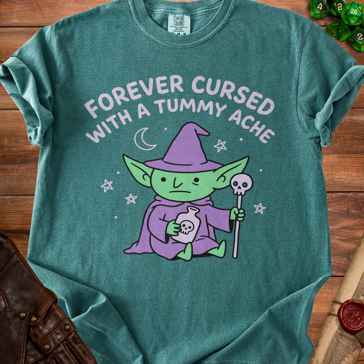 Green t-shirt with a cartoon goblin wearing a witch hat, holding a potion and staff. "Forever Cursed With A Tummy Ache" text.