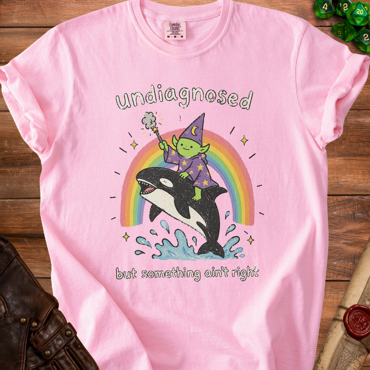 Pink t-shirt with wizard goblin riding an orca on a rainbow, text "undiagnosed but something ain't right".