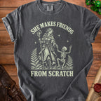 Dark grey t-shirt with graphic of woman and skeleton, text "SHE MAKES FRIENDS FROM SCRATCH".