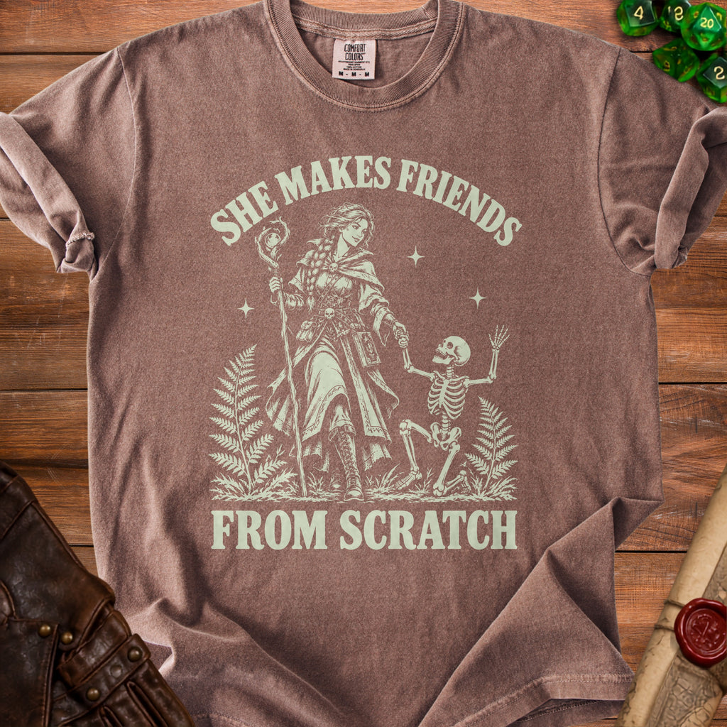 Brown t-shirt with a graphic of a wizard and skeleton, text "She Makes Friends From Scratch".
