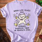 Light purple t-shirt with illustration of a cat squeezing lemons and text "When life gives you lemons, squeeze them in people's eyes."