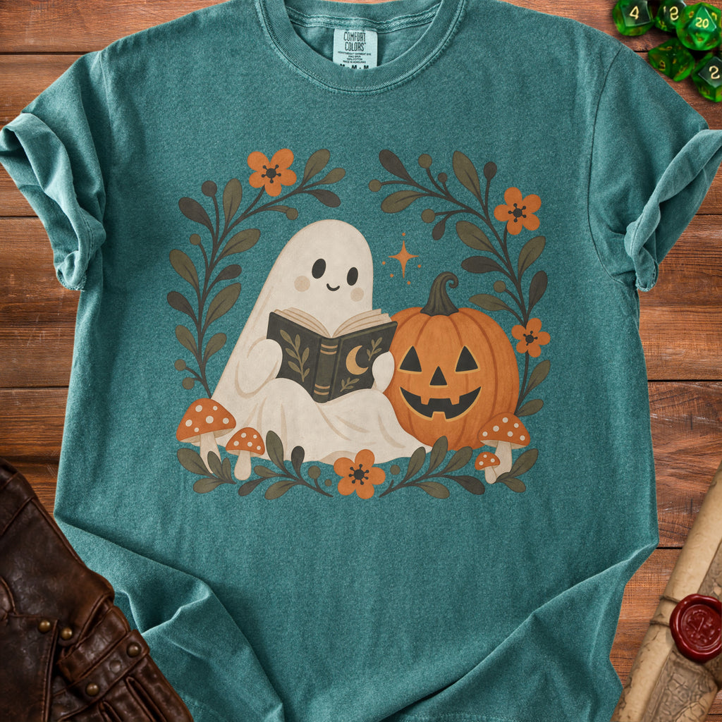 Teal t-shirt featuring a cartoon ghost reading a book beside a jack-o'-lantern, framed by autumn foliage and mushrooms.