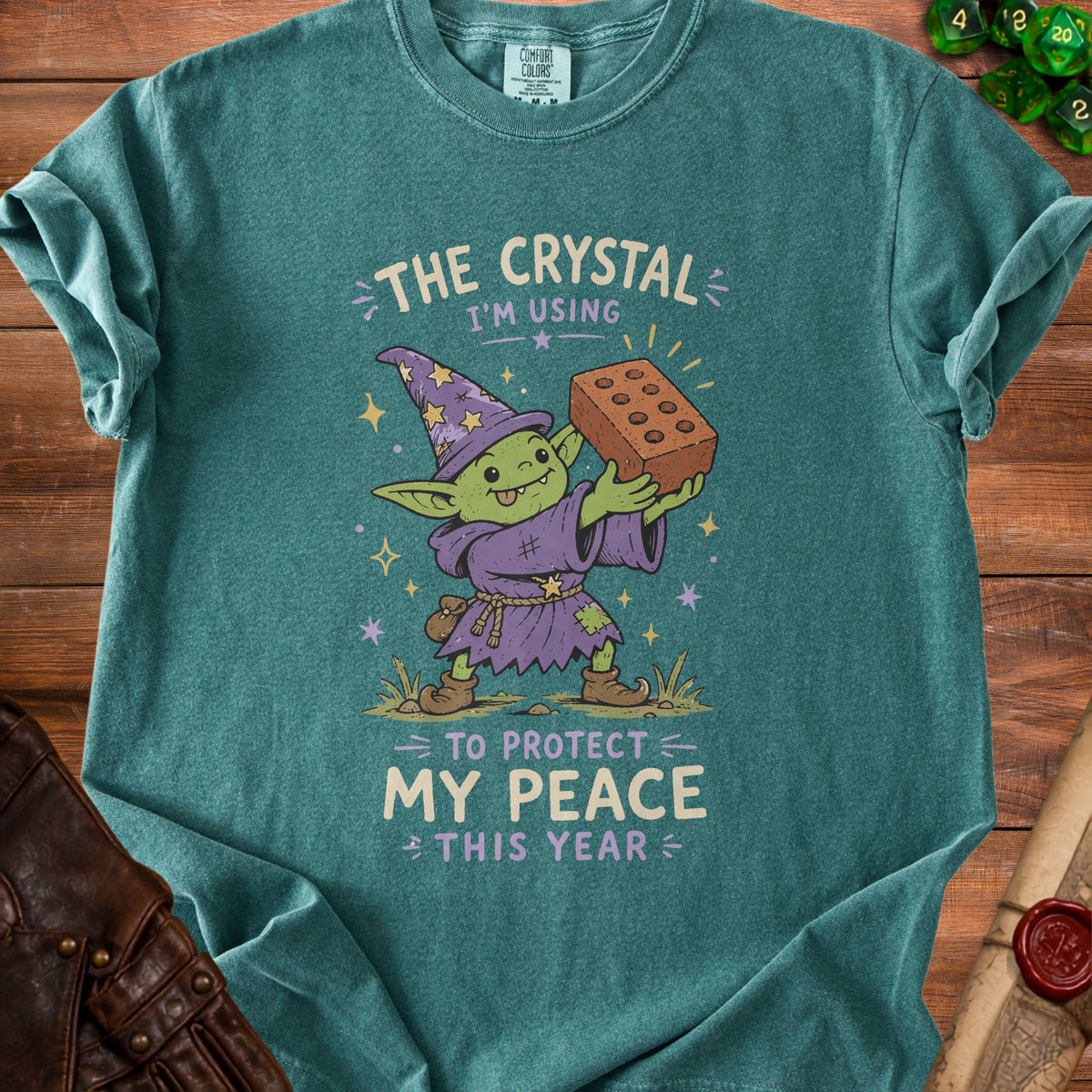 Teal t-shirt with illustration of a goblin wizard holding a brick, text reads "The crystal I'm using to protect my peace this year".