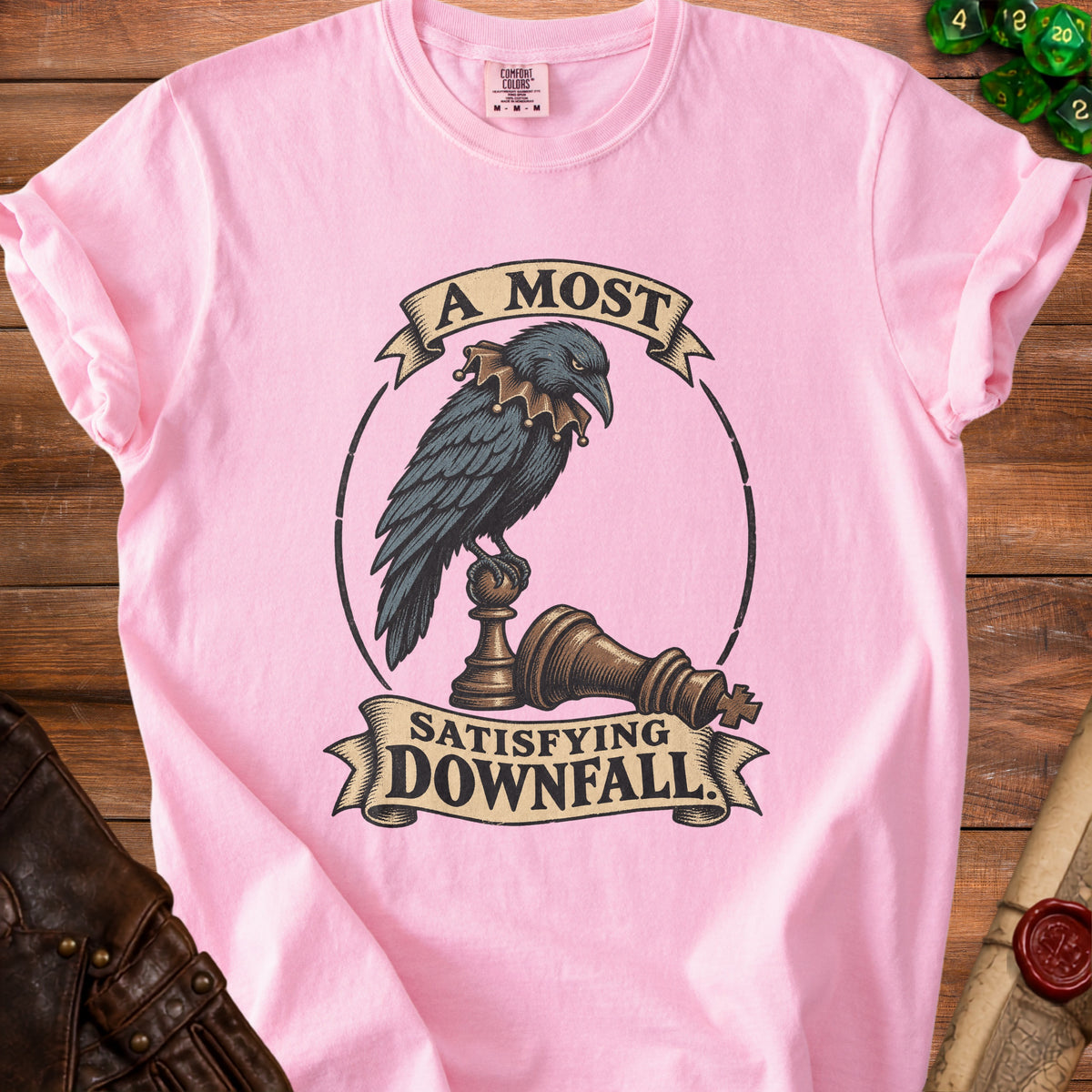 A pink t-shirt with a graphic of a crow perched on a fallen chess piece and the text A Most Satisfying Downfall.