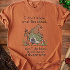 Orange t-shirt with graphic of a mushroom house character and text "I don't know what lies ahead... but I do know it will be an adventure".