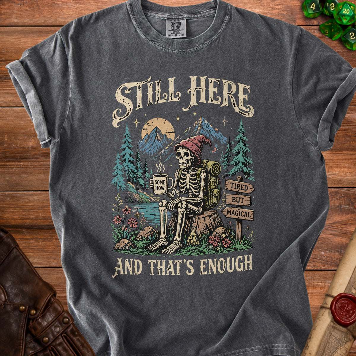 Skeleton sitting with coffee amidst mountains, reading "Still Here, Tired But Magical."