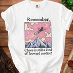 White t-shirt with a graphic of a black cat flying on a broomstick over mountains, reading "Remember, Chaos is still a kind of forward motion!"