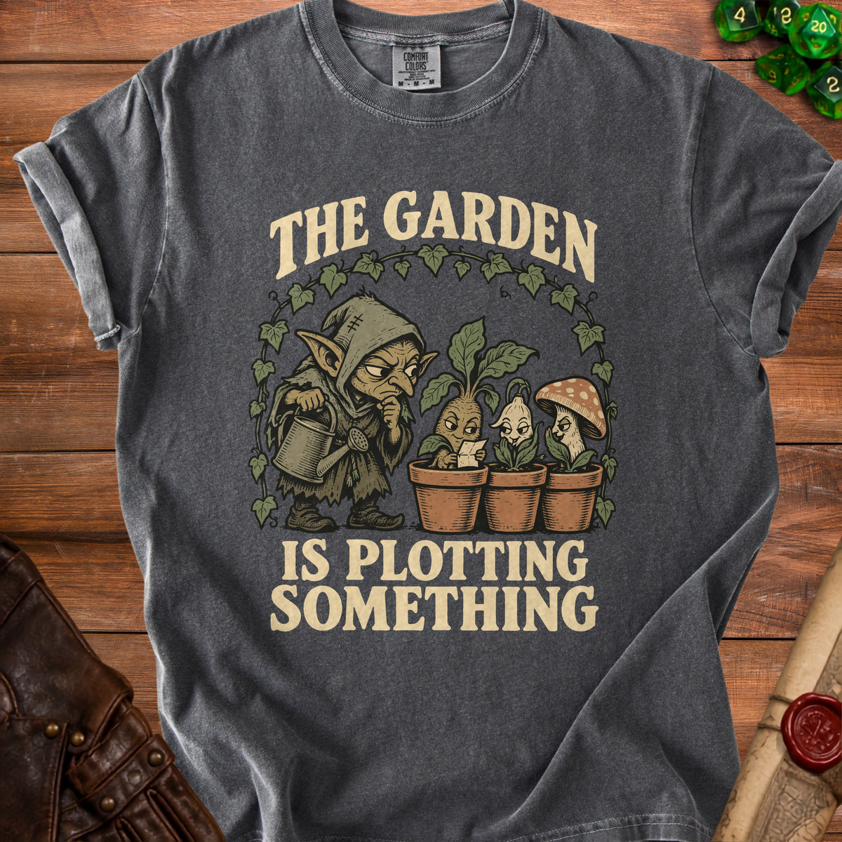 T-shirt with a goblin watering plants and the text "The Garden Is Plotting Something".