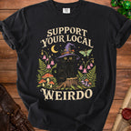 A black t-shirt featuring a graphic of a black cat in a witch hat surrounded by mushrooms, flowers, and the text Support Your Local Weirdo.