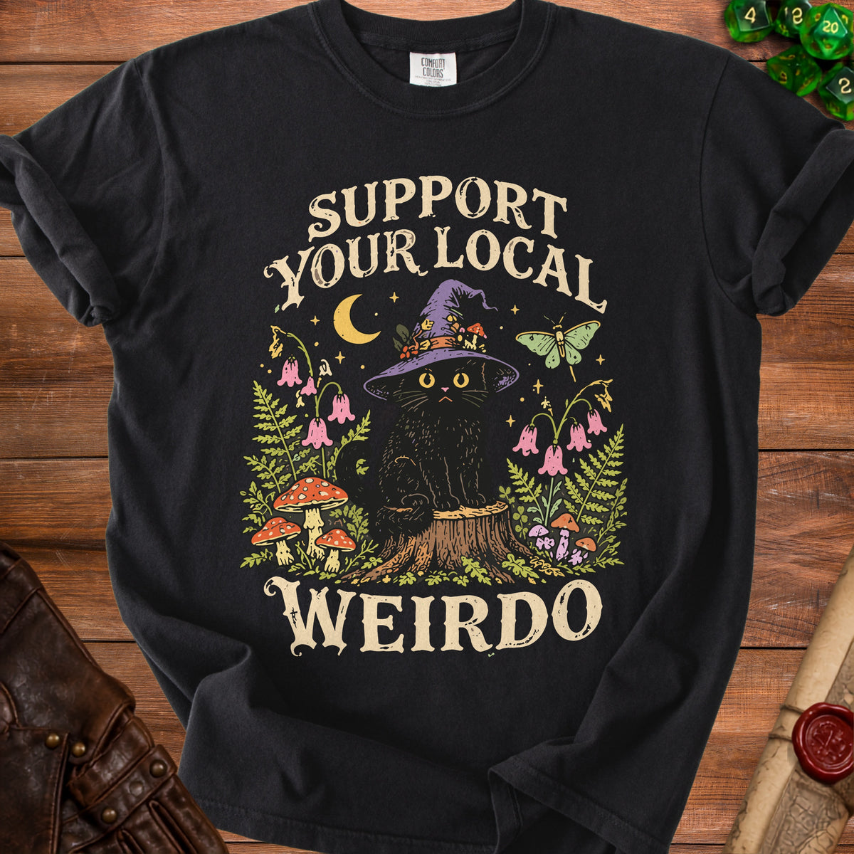 A black t-shirt featuring a graphic of a black cat in a witch hat surrounded by mushrooms, flowers, and the text Support Your Local Weirdo.