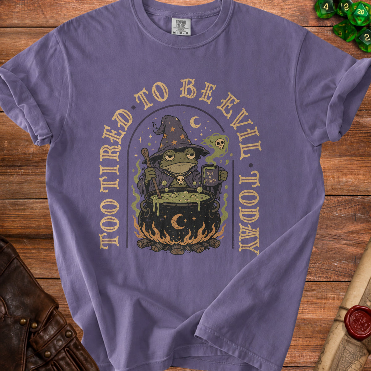 Purple t-shirt with a graphic of a wizard frog by a cauldron, text reads "Too tired to be evil today".