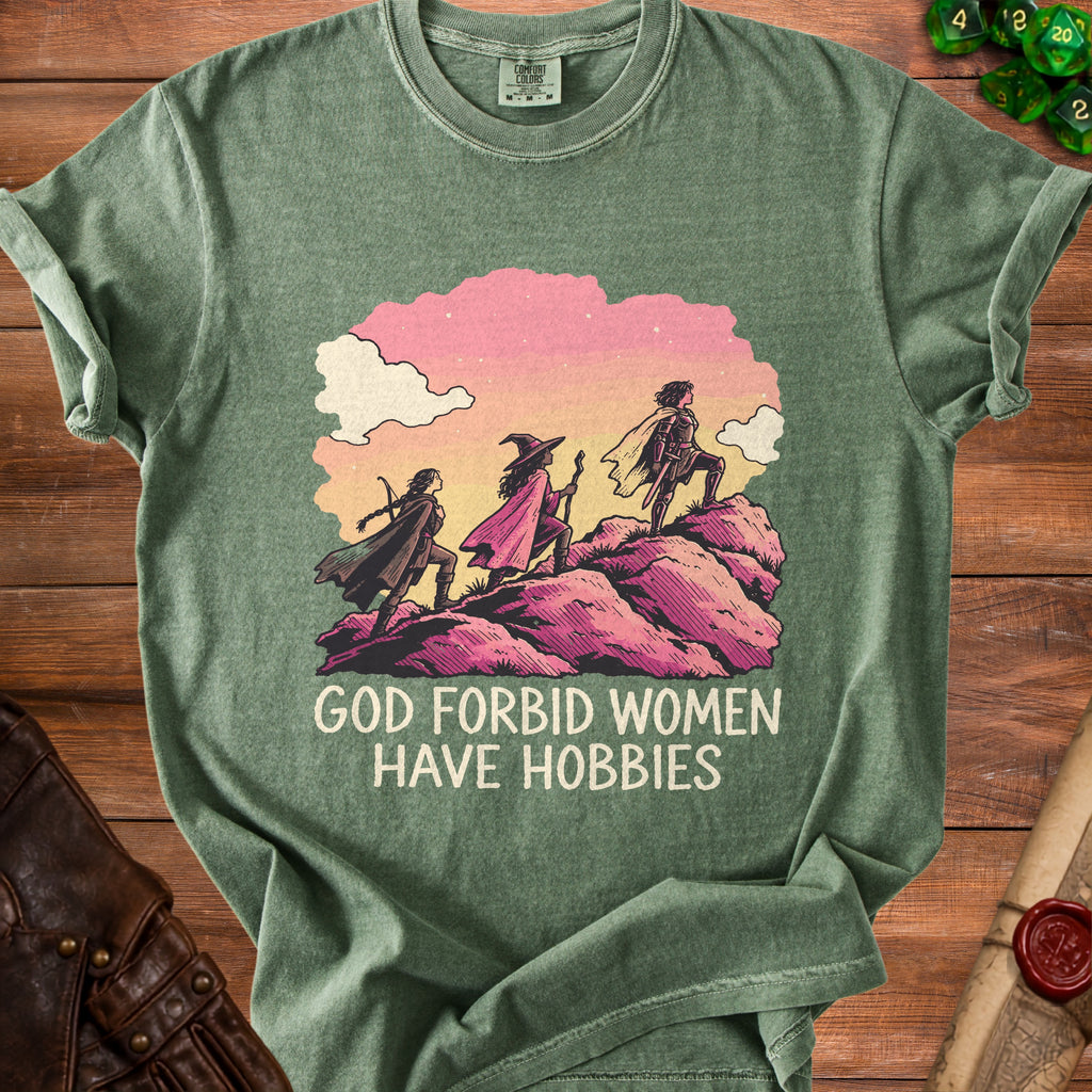 Sage green t-shirt featuring three fantasy women climbing a rocky hill, with text reading GOD FORBID WOMEN HAVE HOBBIES.
