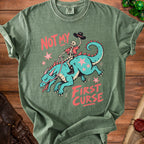 Green t-shirt with skeleton cowboy riding a teal dragon and text "NOT MY FIRST CURSE".