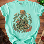 Mint t-shirt with a bottle illustration containing a D20 die and forest elements.