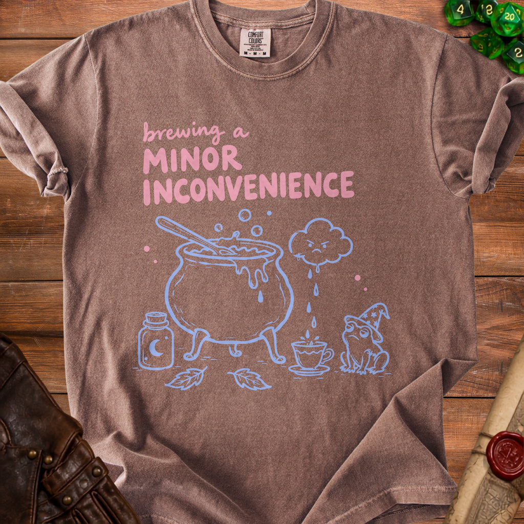 Brown t-shirt with "Brewing a Minor Inconvenience" graphic, cauldron, frog, and dice.