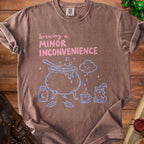 Brown t-shirt with "Brewing a Minor Inconvenience" graphic, cauldron, frog, and dice.