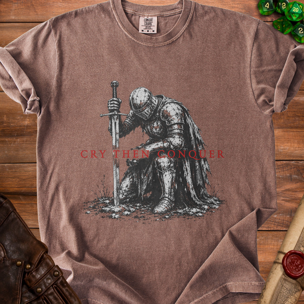 Brown t-shirt graphic of a kneeling knight with sword and "Cry Then Conquer" text.