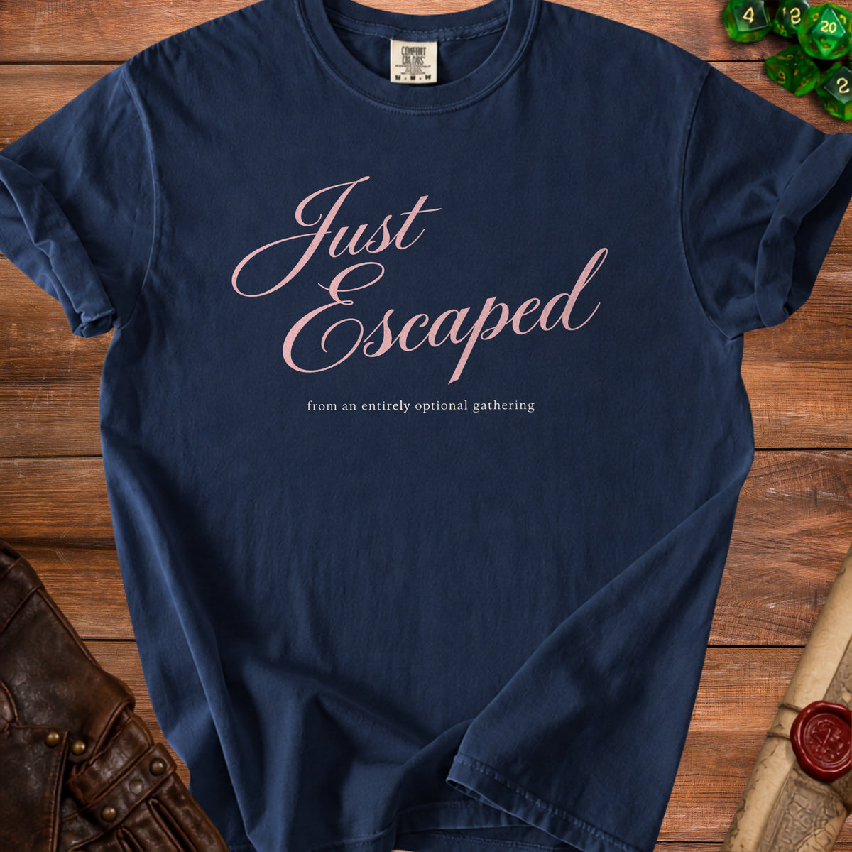 Dark blue t-shirt with pink cursive text reading Just Escaped from an entirely optional gathering on wooden table.