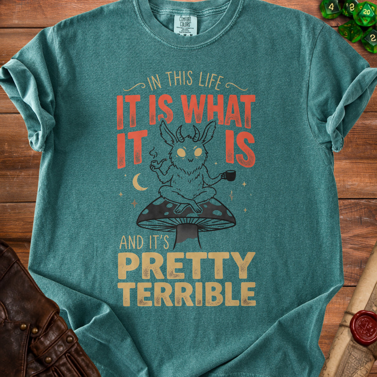 Teal Comfort Colors shirt with goat-like creature sitting on mushroom, text reads "In this life it is what it is and it's pretty terrible."