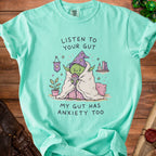 Mint green t-shirt with illustration of anxious goblin holding "Worry Juice". Text reads: Listen to your gut, my gut has anxiety too.